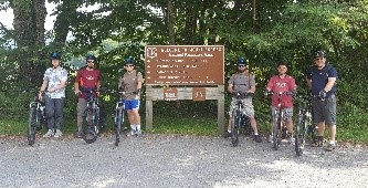Group of cyclists posing with their bikes beside a trailhead sign in a wooded area.