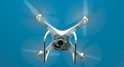 Quadcopter drone hovering against a blue sky, with spinning rotors blurred in motion.