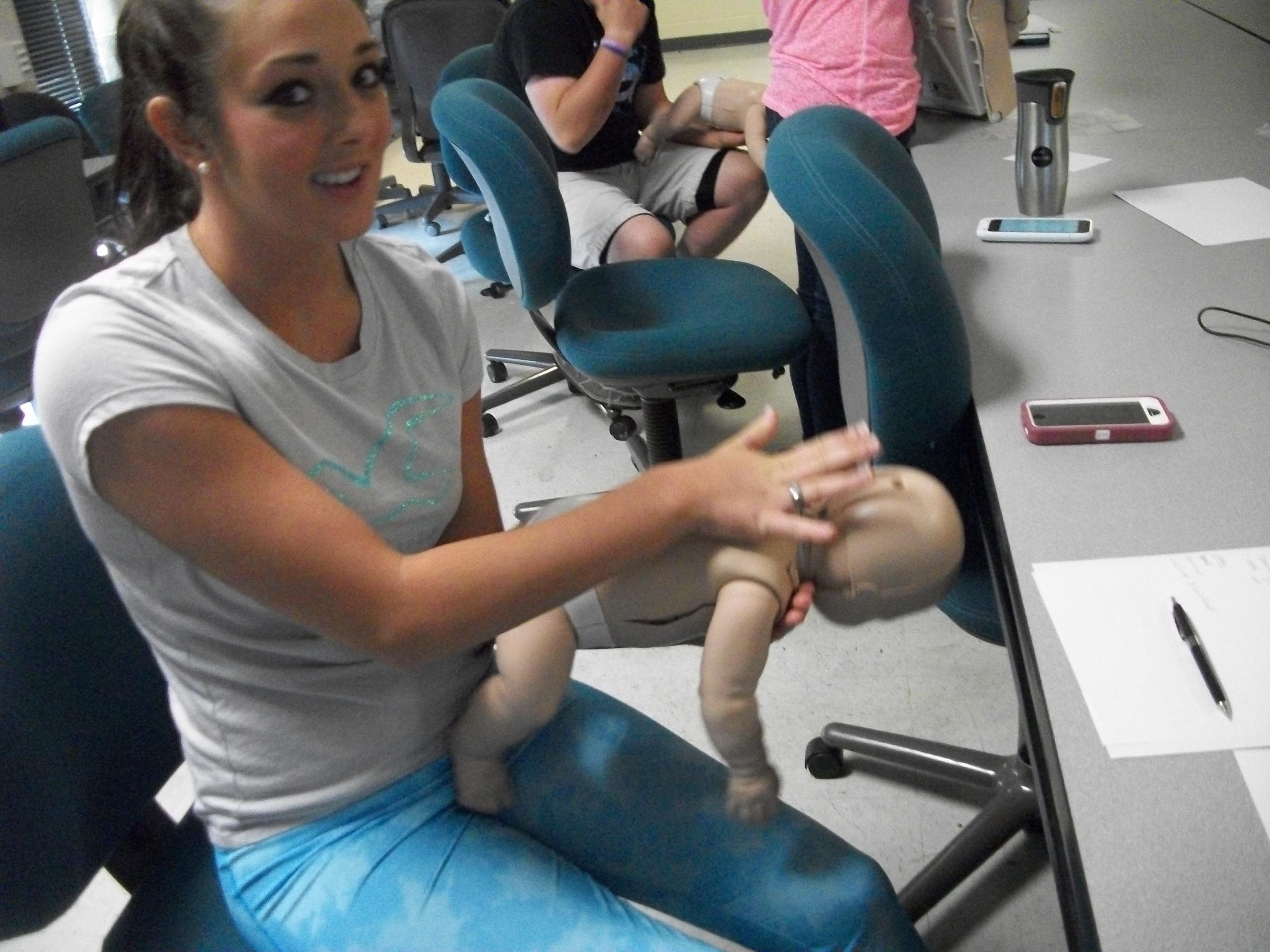 Person in a classroom setting practices infant CPR, holding a baby mannequin over their lap while other students sit in the background.