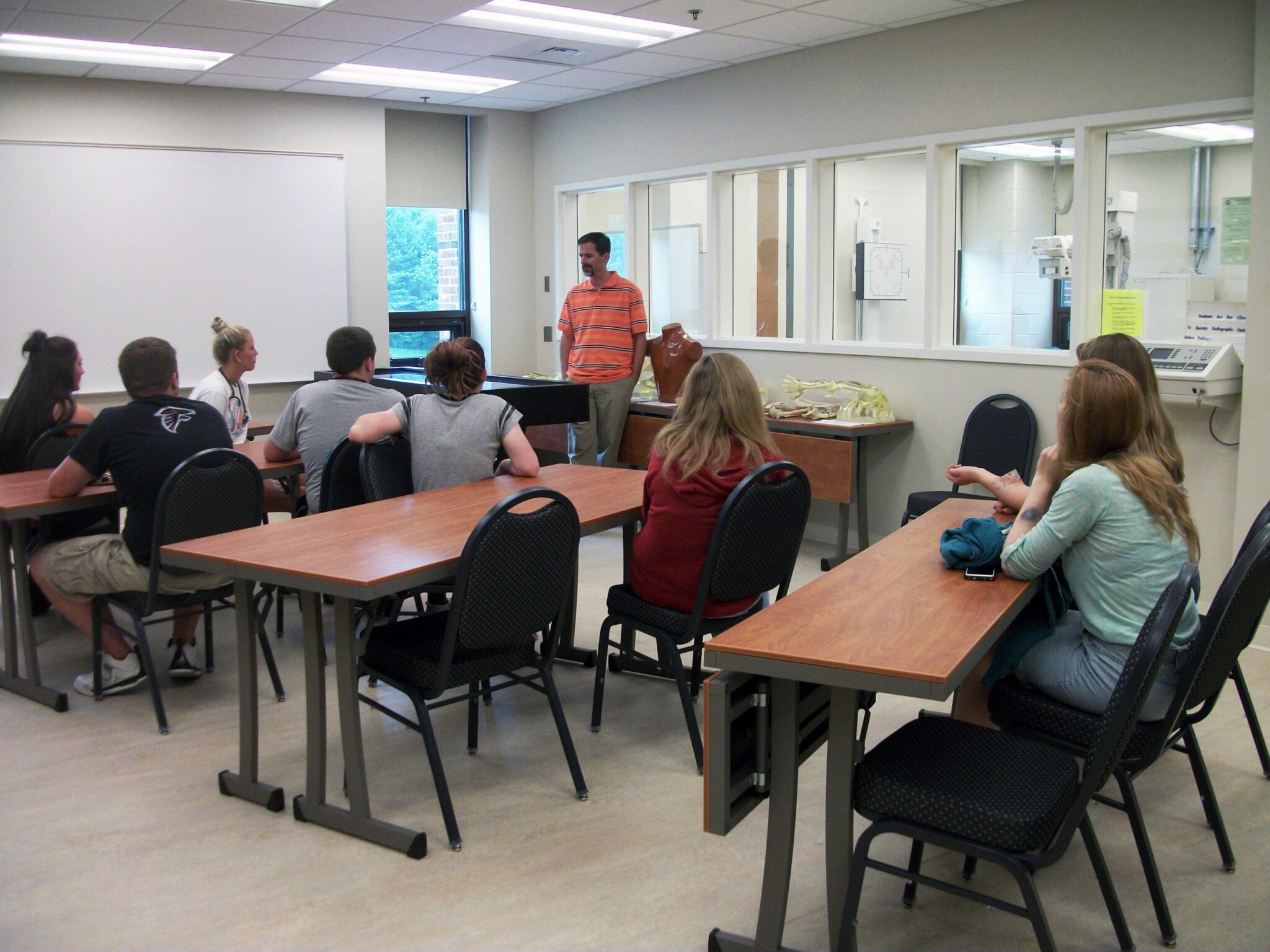 Instructor stands at the front of a classroom speaking to seated students at tables, with a lab observation window and medical training equipment in the background.