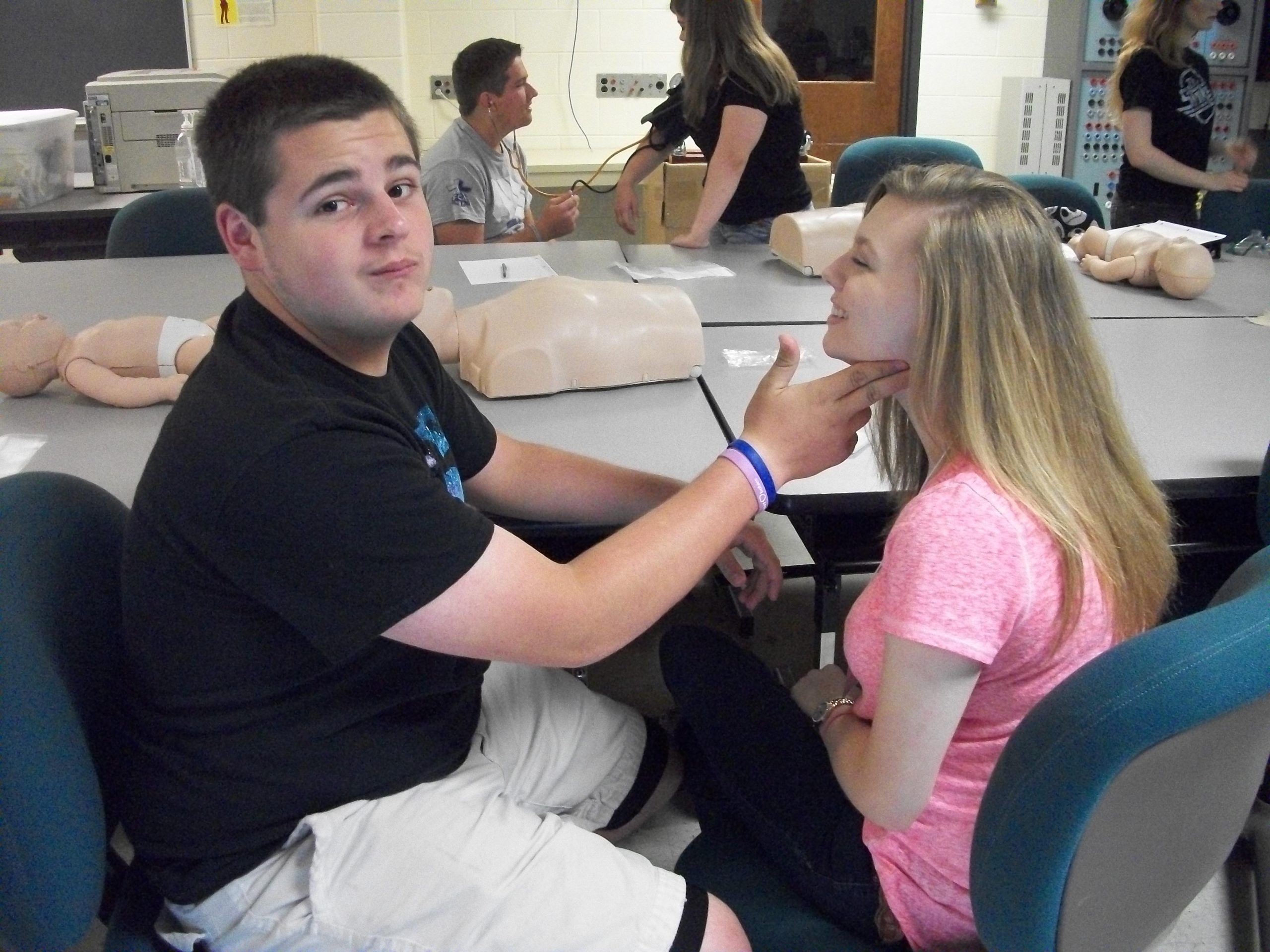 Students practice a medical assessment in a classroom lab, with one student checking another’s neck while CPR training mannequins sit on the tables.