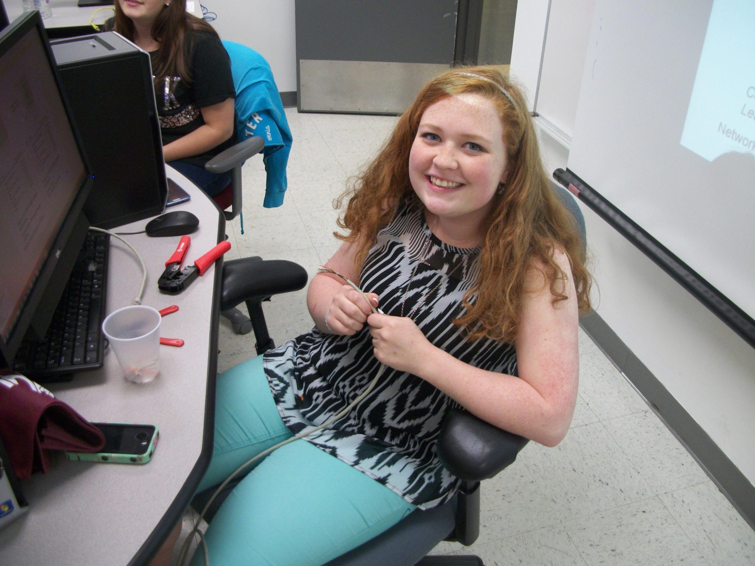 Student smiling while holding an Ethernet cable at a computer lab workstation, with monitors and crimping tools on the desk.