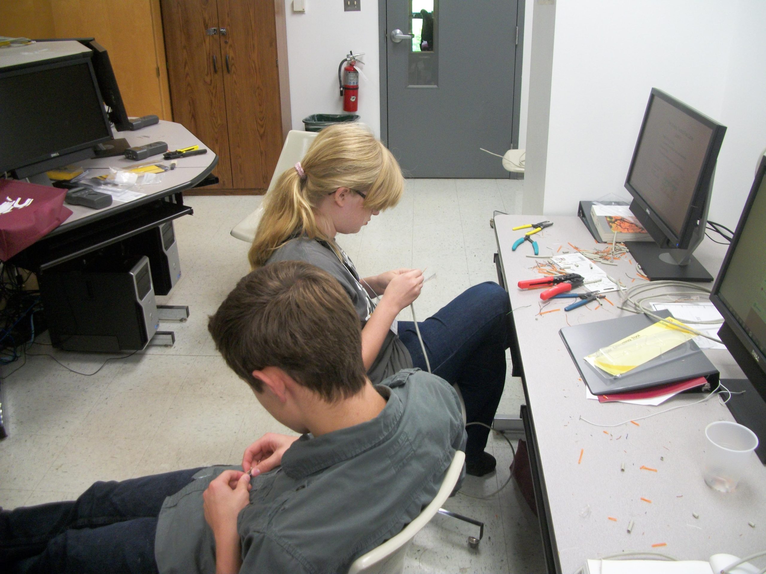 Two students in a computer lab work with wires and hand tools at a cluttered desk beside computer monitors.
