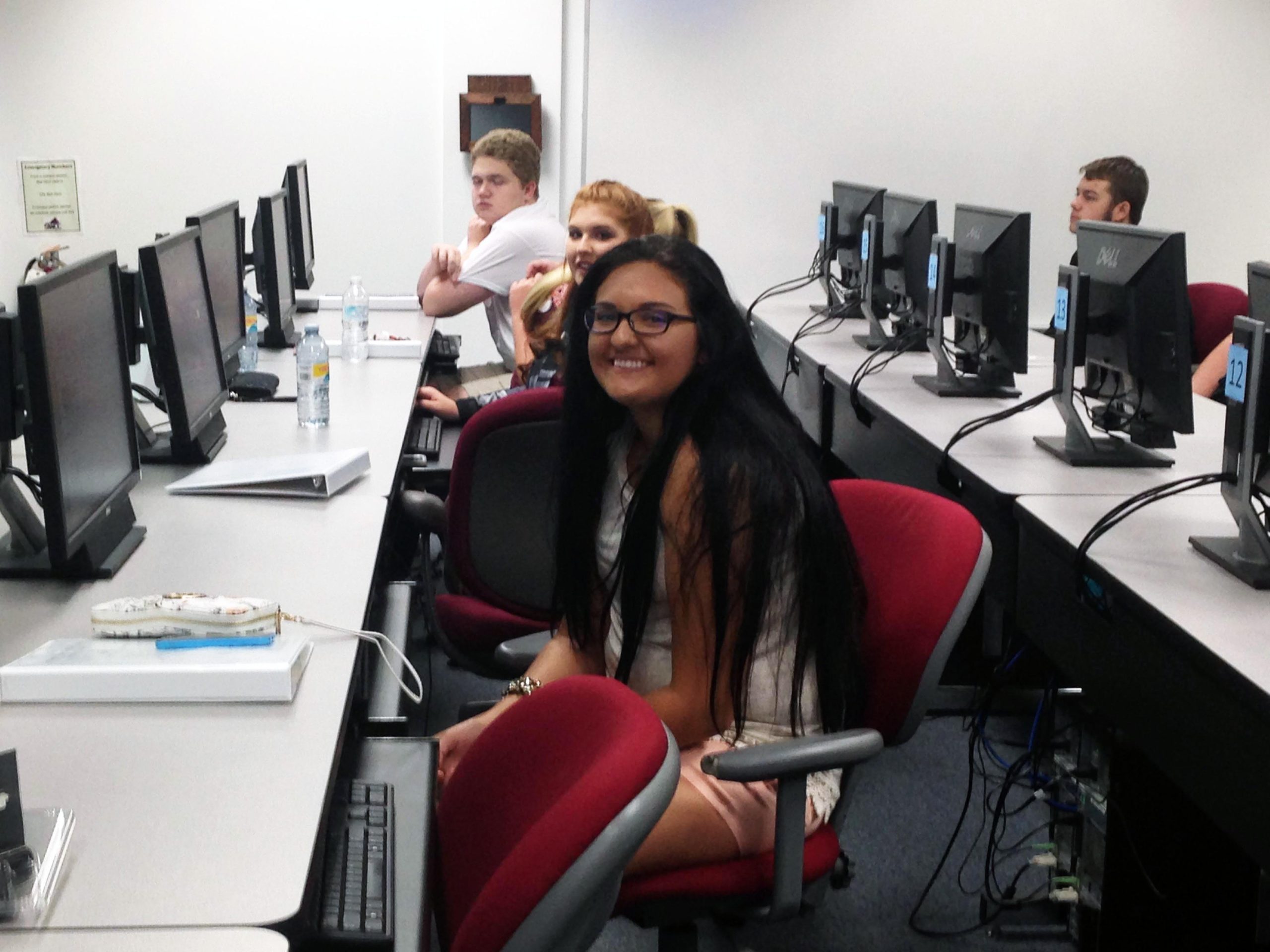 Students seated in a computer lab with rows of monitors; one student in the foreground smiles toward the camera.