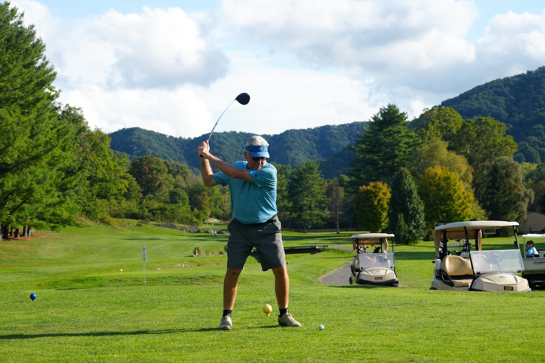 Golfer prepares to tee off on a mountain-lined course with golf carts and trees in the background.