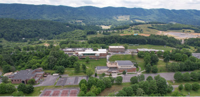 Aerial view of a college campus surrounded by rolling green hills and forested mountains. Multiple academic buildings with brick and light-colored roofs are connected by sidewalks and roads, with parking areas, tennis courts, and open grassy spaces visible in the foreground. The campus is nestled among trees, with scattered farmhouses and fields extending into the hillside landscape under a partly cloudy sky.