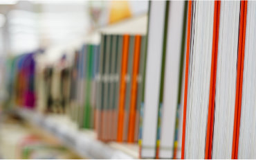 Close-up view of books arranged upright on a shelf, showing colorful spines with white page edges in sharp focus. The books extend into the background with a shallow depth of field, creating a blurred effect that suggests a bookstore or library setting under bright indoor lighting.