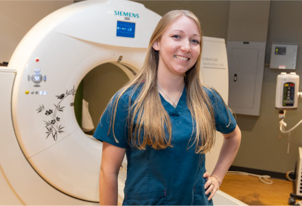 A person wearing teal medical scrubs stands in front of a large Siemens computed tomography (CT) scanner in a clinical imaging room. The CT scanner’s circular opening is visible behind the person, along with control panels, medical equipment, and cabinets in a well‑lit healthcare setting.