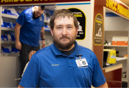 Person standing in the foreground of an emergency medical services training lab, wearing a blue polo shirt with a visible “Student” name badge and an ID card clipped to the chest. In the background, an ambulance training bay is visible with another person inside the open ambulance and shelves of medical equipment. The setting includes bright indoor lighting, instructional signage, and simulated emergency response equipment.