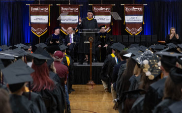 Wide-angle view of a graduation ceremony inside a gymnasium or auditorium. In the foreground, rows of seated graduates wear black caps and gowns, facing a raised stage. At the center of the stage, a speaker stands behind a lectern while several other individuals in academic regalia sit nearby. Behind the stage, four vertical banners hang against dark blue curtains, each banner reading “Southwest” at the top with additional text below. The scene is lit with warm stage lighting, and a person at the far right of the stage appears to be providing sign language interpretation.