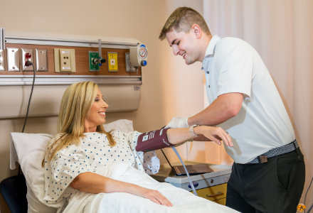 A healthcare setting shows a patient reclining in a hospital bed while a medical professional places a blood‑pressure cuff on the patient’s arm. The room includes wall‑mounted medical equipment such as oxygen outlets, monitoring ports, and electrical panels above the bed.