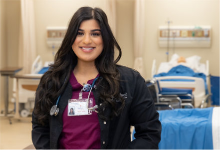 Person standing in the foreground of a healthcare training classroom, wearing burgundy medical scrubs under a black jacket with an ID badge and stethoscope visible. Behind the person are hospital beds with blue linens, bedside equipment, and wall-mounted medical fixtures, arranged in a bright, simulated clinical learning environment.