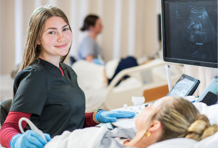 Person wearing black medical scrubs and blue gloves performs an ultrasound scan on a patient lying on a hospital bed. An ultrasound transducer is held against the patient’s abdomen, and a nearby monitor displays a grayscale ultrasound image. The setting is a bright clinical training environment with hospital beds, medical equipment, and another bed visible in the background.