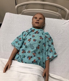 Pediatric medical training mannequin lying on a hospital bed with head elevated on a pillow and arms at the sides. The mannequin is wearing a light blue hospital gown with a small multicolored pattern and is partially covered by a white blanket. A metal bed rail is visible behind the head in a clinical simulation setting.