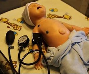 Neonatal medical training mannequin lying on a padded surface with a light blue towel covering the lower body and a cap on the head. A blood pressure cuff is wrapped around the upper arm and connected to a manual bulb pump and gauge resting beside the mannequin. The setting appears to be a clinical simulation environment focused on newborn assessment.