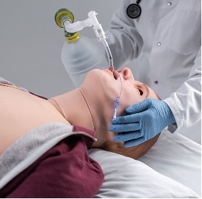 A medical training mannequin lying on a hospital bed while a clinician wearing blue gloves uses a bag-valve mask to ventilate the mannequin through a clear face mask. The clinician is dressed in a white lab coat with a stethoscope visible, and the manual resuscitator bag is connected to the mask by clear tubing in a clinical simulation setting.