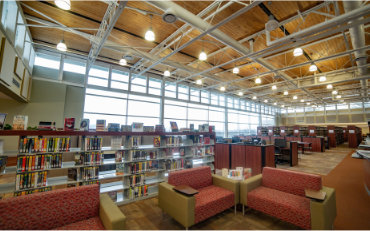Wide-angle view of a modern academic library interior with high ceilings, exposed wooden beams, and large windows along one wall allowing natural light to fill the space. Multiple rows of bookshelves are arranged throughout the room, with seating areas in the foreground featuring red patterned lounge chairs and small tables. The floor is carpeted, overhead lights are evenly spaced, and the space appears open, quiet, and designed for study and reading.