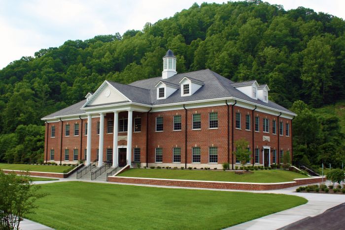 Two-story red-brick building with white columns and a cupola, set against a forested hillside.